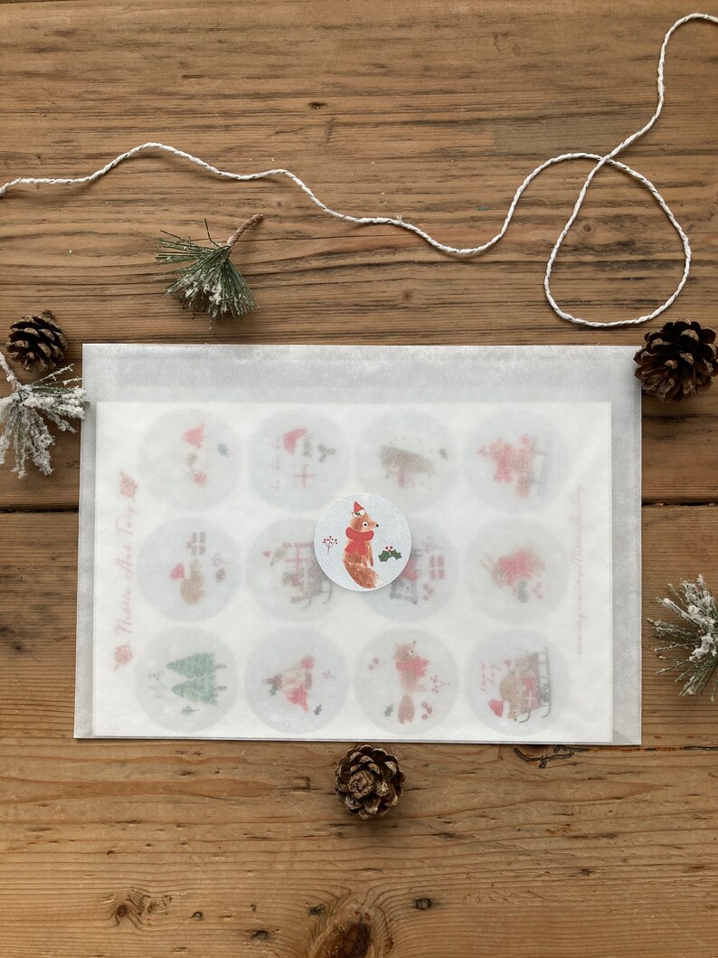 May include: A white envelope with a sheet of paper inside. The paper has 12 round stickers with various Christmas-themed illustrations. The stickers feature a fox, a Christmas tree, a house, and other wintery designs. The text "Merry little Tiny Fox" is printed on the paper.