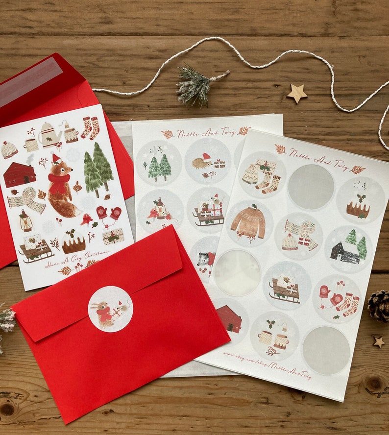 May include: Two sheets of stickers featuring a winter theme with illustrations of a fox, a hedgehog, a house, a sled, mittens, and a teapot. The stickers are on a white background with a gray border. The text "Nibble And Twig" is printed on the sheets. The stickers are packaged in a red envelope with the text "Have a Cozy Christmas" printed on the front.