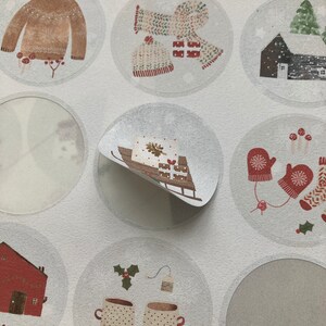 May include: A sheet of round stickers with various winter-themed illustrations. The stickers feature images of a sweater, mittens, a house, a sled, a Christmas tree, and mugs of hot cocoa.