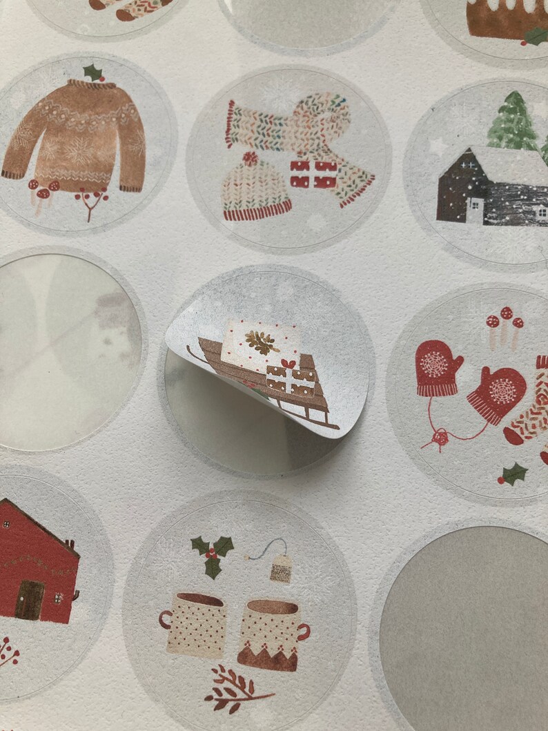 May include: A sheet of round stickers with various winter-themed illustrations. The stickers feature images of a sweater, mittens, a house, a sled, a Christmas tree, and mugs of hot cocoa.