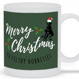 May include: White ceramic mug with a green background featuring the text "Merry Christmas Ya Filthy Hobbitsees" in white script. A black silhouette of a creature wearing a Santa hat is also on the mug. Holly accents are included.