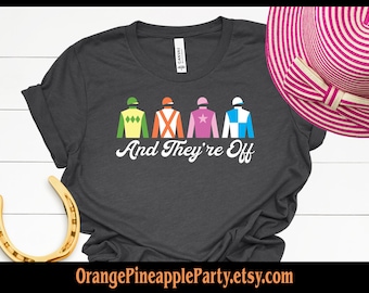 May include: A dark gray t-shirt with a white graphic of four jockeys in different colored silks. The text "And They're Off" is written below the jockeys.