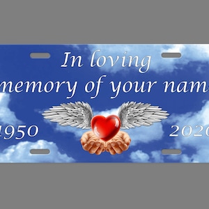 May include: A blue and white memorial license plate with a white heart with wings and the text "In loving memory of your name 1950 2020".