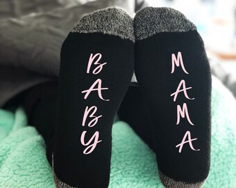 May include: A pair of black socks with gray trim. One sock has "BABY" written on it in pink letters, and the other sock has "MAMA" written on it in pink letters. The socks are on a light green surface.