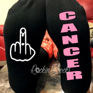 以下が含まれることがあります： 片方の靴下に白い中指、もう片方の靴下にピンクの文字で「CANCER」と書かれた黒い靴下1足。靴下は赤いソファを背景にテーブルの上に置かれている。