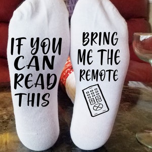 May include: A pair of white socks with black text. One sock says "If you can read this" and the other sock says "Bring me the remote" with a black and white illustration of a TV remote.