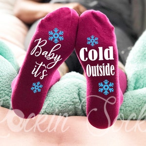 May include: A pair of burgundy socks with white text. One sock says "Baby it's" and the other says "Cold Outside". Both socks have a blue snowflake design.