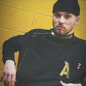 Op de afbeelding: Een persoon draagt een zwart koksjack met gouden details en een zwarte beanie. Het jack heeft een gouden "A" op de zak en de tekst "FADESBYFISH" op de kraag.