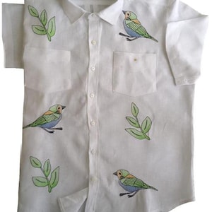 May include: White short-sleeved button-down shirt with green leaves and colorful birds embroidered on the front. The shirt has a pocket on the left side and the text "Isla de Mar" on the collar.