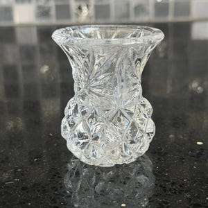 May include: Clear glass bud vase with a thistle-like design. The vase features a flared top and a textured, diamond-patterned base. The glass reflects light, creating a sparkling effect. The vase is likely for displaying small flowers or stems.