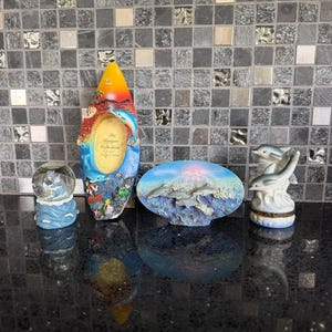 May include: A collection of dolphin-themed decorative items. Includes a snow globe, a photo frame with a dolphin design, an oval-shaped plaque with dolphins, and a small ceramic figurine of dolphins. The items are in shades of blue, white, and yellow.