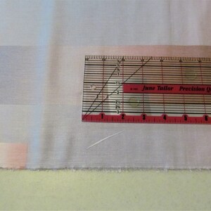 May include: A close-up of a white fabric with a pink and blue ombre design. A red and white quilting ruler with the text "June Tailor Precision Quilting Rule" is placed on top of the fabric.