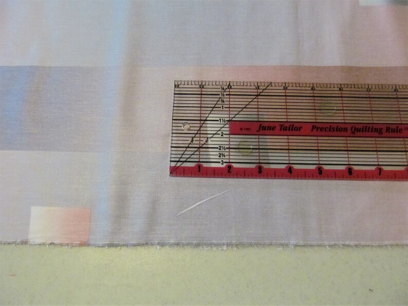 May include: A close-up of a white fabric with a pink and blue ombre design. A red and white quilting ruler with the text "June Tailor Precision Quilting Rule" is placed on top of the fabric.
