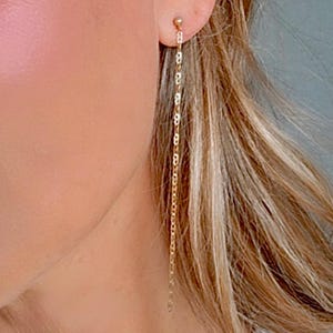 May include: A close-up of a gold chain earring. The earring is a long, delicate chain that hangs down from the earlobe. The earring is a simple, elegant design.