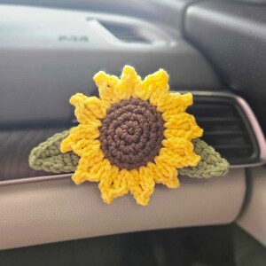 May include: A crocheted sunflower car air vent clip. The sunflower has yellow petals, a brown center, and green leaves. The handmade accessory is designed to clip onto a car's air vent.