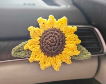 Op de afbeelding: Een gehaakte zonnebloem auto ventilatieclip. De zonnebloem heeft gele bloemblaadjes, een bruin midden en groene bladeren. Het handgemaakte accessoire is ontworpen om op een autoventilatie te klikken.
