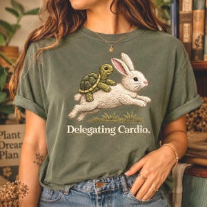 May include: An olive green t-shirt featuring a graphic of a small green turtle riding on the back of a white rabbit. Below the illustration, the text reads "Delegating Cardio." The shirt has short sleeves and a crew neckline, made of cotton.