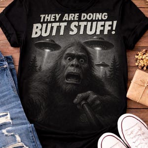 May include: Black t-shirt with the text "THEY ARE DOING BUTT STUFF!" above a graphic of a surprised Bigfoot being illuminated by three UFOs. The shirt is displayed with blue jeans and white sneakers on a wooden surface.