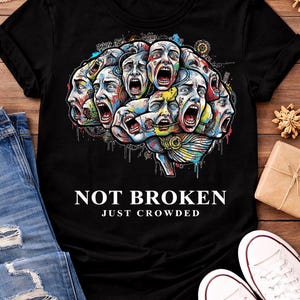 May include: Black t-shirt featuring a colorful graphic of multiple screaming faces arranged in a brain shape. The text "NOT BROKEN JUST CROWDED" is printed below the graphic. The shirt is paired with blue jeans and white sneakers.