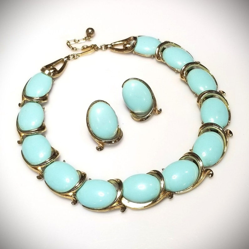 May include: A vintage jewelry set featuring a necklace and matching earrings. The necklace and earrings have oval, light blue cabochons set in gold-tone metal. The necklace is adjustable and the earrings are clip-ons. The set is displayed on a white background.