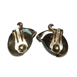 May include: A pair of vintage clip-on earrings. Each earring features a light blue oval stone set in a gold-toned metal frame. The clip mechanism is visible on the back, with the word "URBANTY" stamped on the clip.