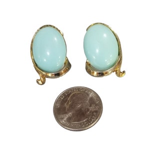 May include: A pair of vintage clip-on earrings featuring oval, light blue cabochons set in gold-tone metal frames. The earrings have a decorative, curved design. A quarter is included for size reference.