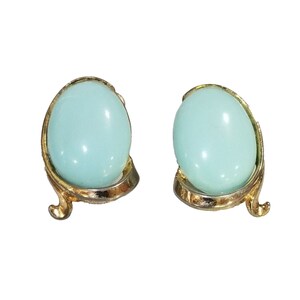 May include: A pair of vintage clip-on earrings. Each earring features a light blue, oval-shaped stone set in a gold-tone metal frame with a decorative, curved design. The earrings have a glossy finish.
