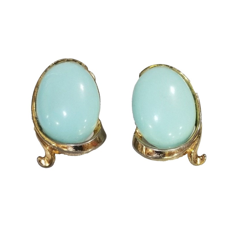 May include: A pair of vintage clip-on earrings. Each earring features a light blue, oval-shaped stone set in a gold-tone metal frame with a decorative, curved design. The earrings have a glossy finish.