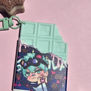 May include: A mint green chocolate bar keychain with a bite taken out, revealing a colorful anime-style illustration of a character holding a heart. The keychain has a green clasp and is set against a pink background.