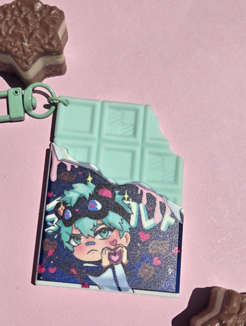 May include: A mint green chocolate bar keychain with a bite taken out, revealing a colorful anime-style illustration of a character holding a heart. The keychain has a green clasp and is set against a pink background.