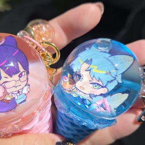 Pode incluir: Dois chaveiros estilo anime. Um chaveiro é rosa e apresenta um personagem de cabelo roxo com óculos segurando um pastel. O outro é azul e apresenta um personagem de cabelo azul. Ambos os chaveiros têm um design transparente em forma de bolha.