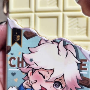 May include: A white chocolate bar with a bite taken out of it, next to a charm with a cartoon character holding a lollipop. The charm has a blue background with the word "CHO" visible. The charm has a gold-colored keychain attachment.