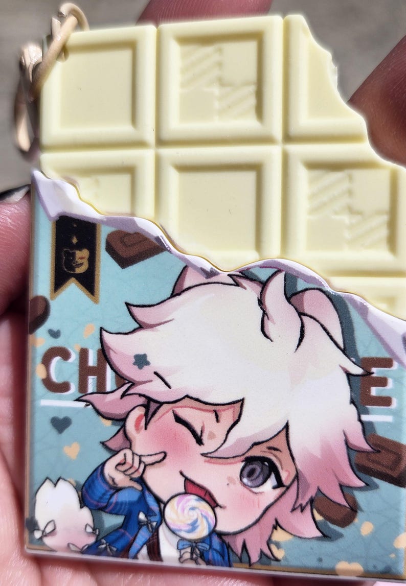 May include: A white chocolate bar with a bite taken out of it, next to a charm with a cartoon character holding a lollipop. The charm has a blue background with the word "CHO" visible. The charm has a gold-colored keychain attachment.
