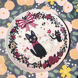 May include: A white ceramic plate featuring a whimsical design with black and white cats, a red and white bow, and a floral border of red roses. The plate is decorated with cookies and pink flower petals.