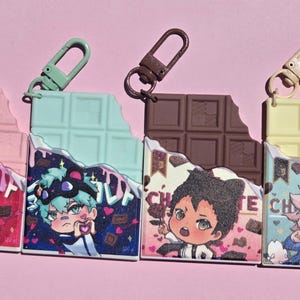 May include: Four colorful anime-style charm keychains shaped like chocolate bars, each with a different color and character design. The keychains have a bite taken out of them and are attached to a metal clasp.
