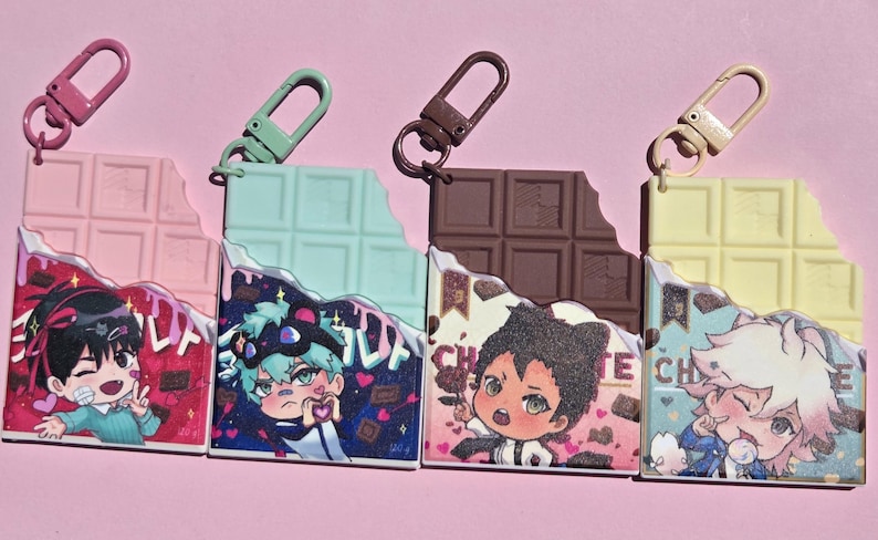 May include: Four colorful anime-style charm keychains shaped like chocolate bars, each with a different color and character design. The keychains have a bite taken out of them and are attached to a metal clasp.