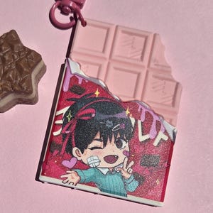 May include: Pink chocolate bar-shaped keychain with a bite taken out, featuring a cartoon character illustration. The keychain has a pink clasp. Two star-shaped chocolates are also visible.