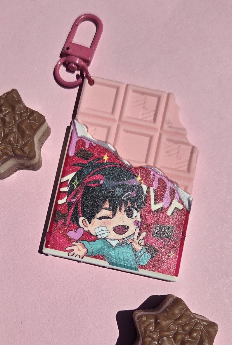 May include: Pink chocolate bar-shaped keychain with a bite taken out, featuring a cartoon character illustration. The keychain has a pink clasp. Two star-shaped chocolates are also visible.