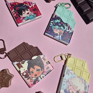 May include: Four novelty keychains shaped like chocolate bars, each with a different color and character design. The keychains have a bite taken out of them, revealing the character art. The background is a light pink color.