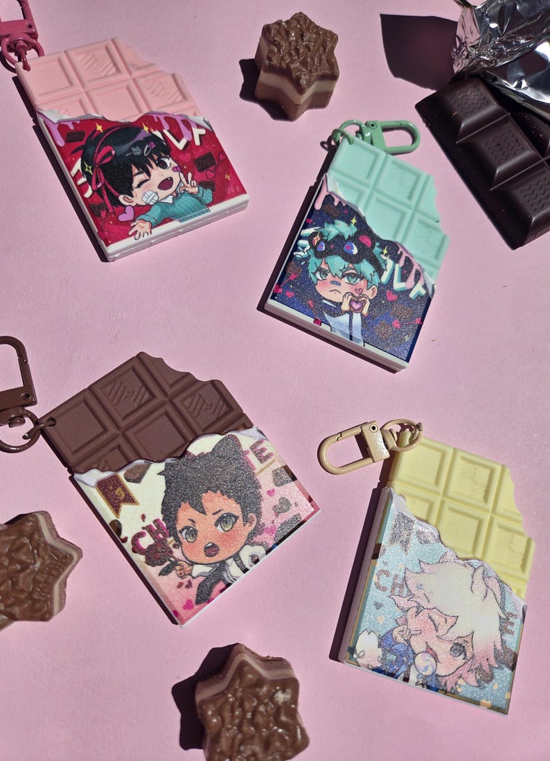 May include: Four novelty keychains shaped like chocolate bars, each with a different color and character design. The keychains have a bite taken out of them, revealing the character art. The background is a light pink color.