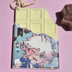 May include: A white chocolate bar-shaped keychain with a bite taken out of it. The keychain has a cartoon image of a character with pink hair and the letters "CH". A small, brown chocolate candy is also visible.