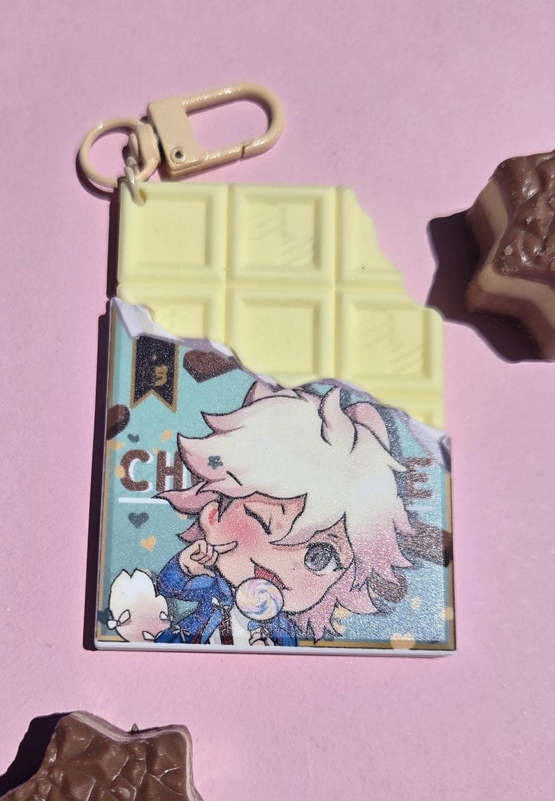 May include: A white chocolate bar-shaped keychain with a bite taken out of it. The keychain has a cartoon image of a character with pink hair and the letters "CH". A small, brown chocolate candy is also visible.
