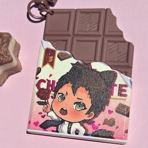May include: A chocolate bar-shaped keychain with a cartoon illustration of a character holding a rose. The keychain has a brown clasp and a bite taken out of the chocolate. The background is pink.