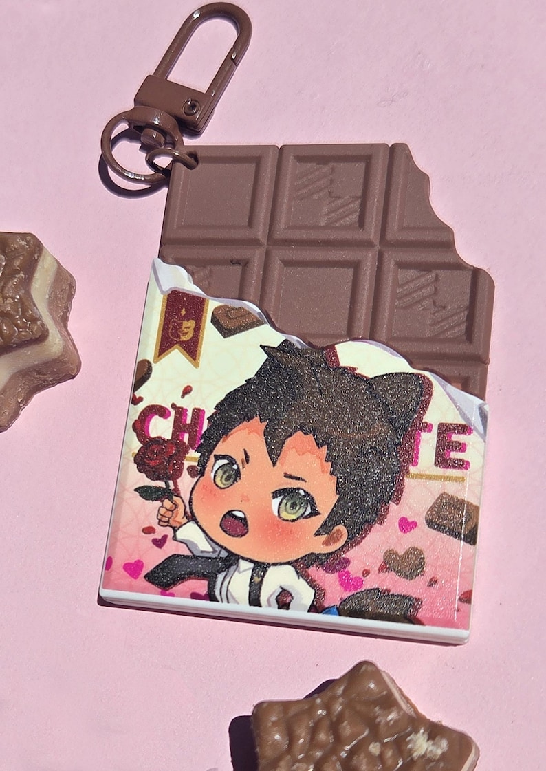 May include: A chocolate bar-shaped keychain with a cartoon illustration of a character holding a rose. The keychain has a brown clasp and a bite taken out of the chocolate. The background is pink.
