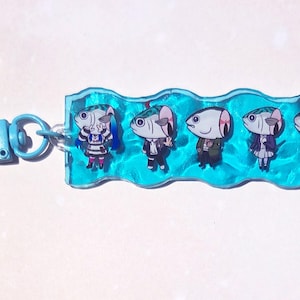 May include: A blue keychain featuring five cartoon fish characters in various outfits. The fish are set against a wavy, turquoise background, with a matching blue clasp.