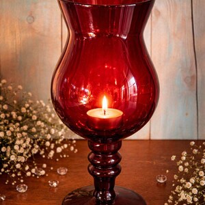 May include: A deep red glass candle holder with a lit tea light inside. The candle holder has a rounded body and a pedestal base. The background features a wooden surface, small white flowers, and clear crystals.