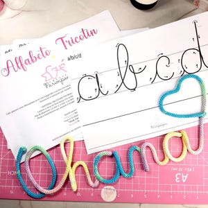 Può includere: Un tappetino da taglio rosa con la parola "Chama" scritta con filo colorato. Le lettere sono in stile corsivo. Ci sono anche fogli stampati con le parole "Alfabeto Tricotin" e le lettere a, b, c e d.