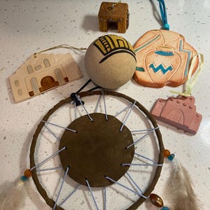 May include: A collection of Southwestern-themed ornaments and a dreamcatcher. The ornaments include a mission-style building, a small wooden box, a round ornament with a geometric design, a pottery-shaped ornament, and a pink building. The dreamcatcher has a brown suede center, blue string, beads, and feathers.