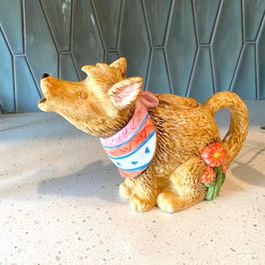 May include: A ceramic teapot shaped like a howling dog with textured brown fur. The dog wears a pink, white, and blue patterned bandana around its neck. A small cluster of orange flowers with green stems is attached to the side of the teapot body.