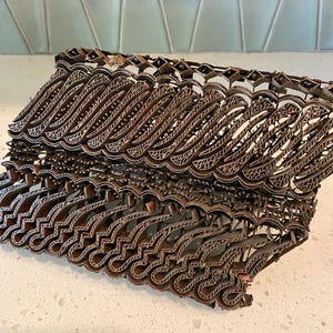 May include: A copper batik tjap tool featuring an intricate, repeating wave-like pattern constructed from thin metal strips. The tool has a structured, rectangular frame with a complex, layered design used for applying wax to fabric in textile arts.